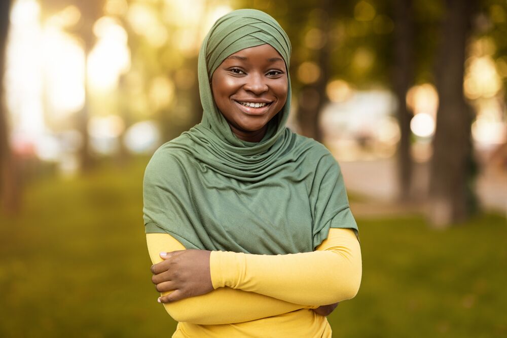 A woman stands in a park and smiles at the camera. She wears a green head covering and a yellow shirt.